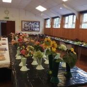A spacious hall with wooden walls featuring long tables filled with various flowers in vases, assorted produce, and baked goods. The room is well-lit with natural light streaming through large windows. Exhibition boards are visible in the background.