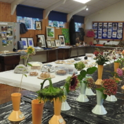 Rows of flowers in vases are displayed on tables alongside baked goods, artwork, and crafts in a community hall with wooden walls and bulletin boards. Two people are seen arranging items in the background.