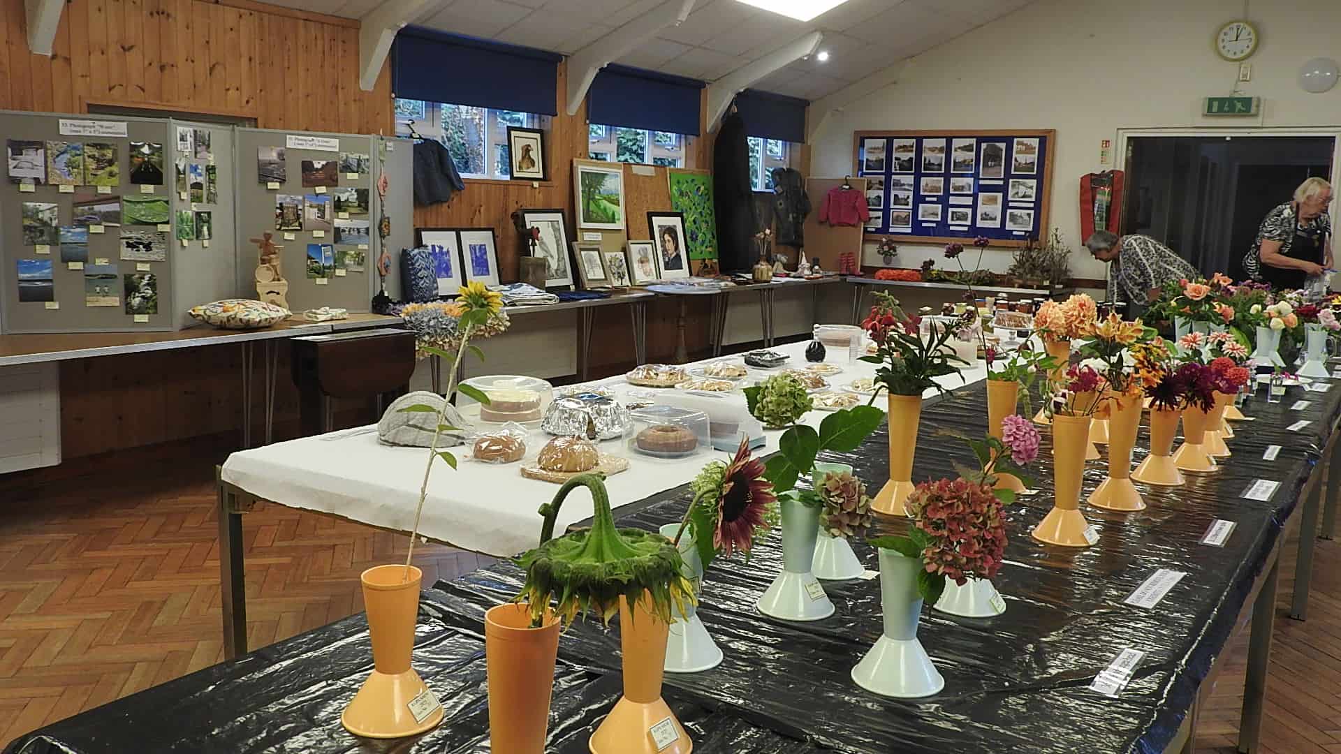 Rows of flowers in vases are displayed on tables alongside baked goods, artwork, and crafts in a community hall with wooden walls and bulletin boards. Two people are seen arranging items in the background.