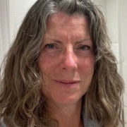 A woman with wavy grey hair and a neutral expression stands indoors in front of a tiled wall, wearing a sleeveless top. A small brown hair clip is visible near her hairline.