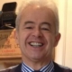 A smiling middle-aged man with short grey hair wearing a blue and white striped shirt and a dark waistcoat, standing indoors with a wooden crate and office equipment in the background.