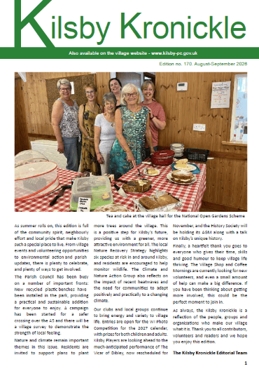A group of people stand behind a table with tea, cake, and cups at a village hall event. The room has posters, a map, and a red and white checked tablecloth on the serving table.